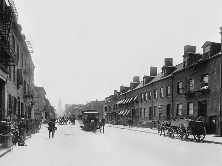 A black and white photo of a street with a horse drawn carriage.